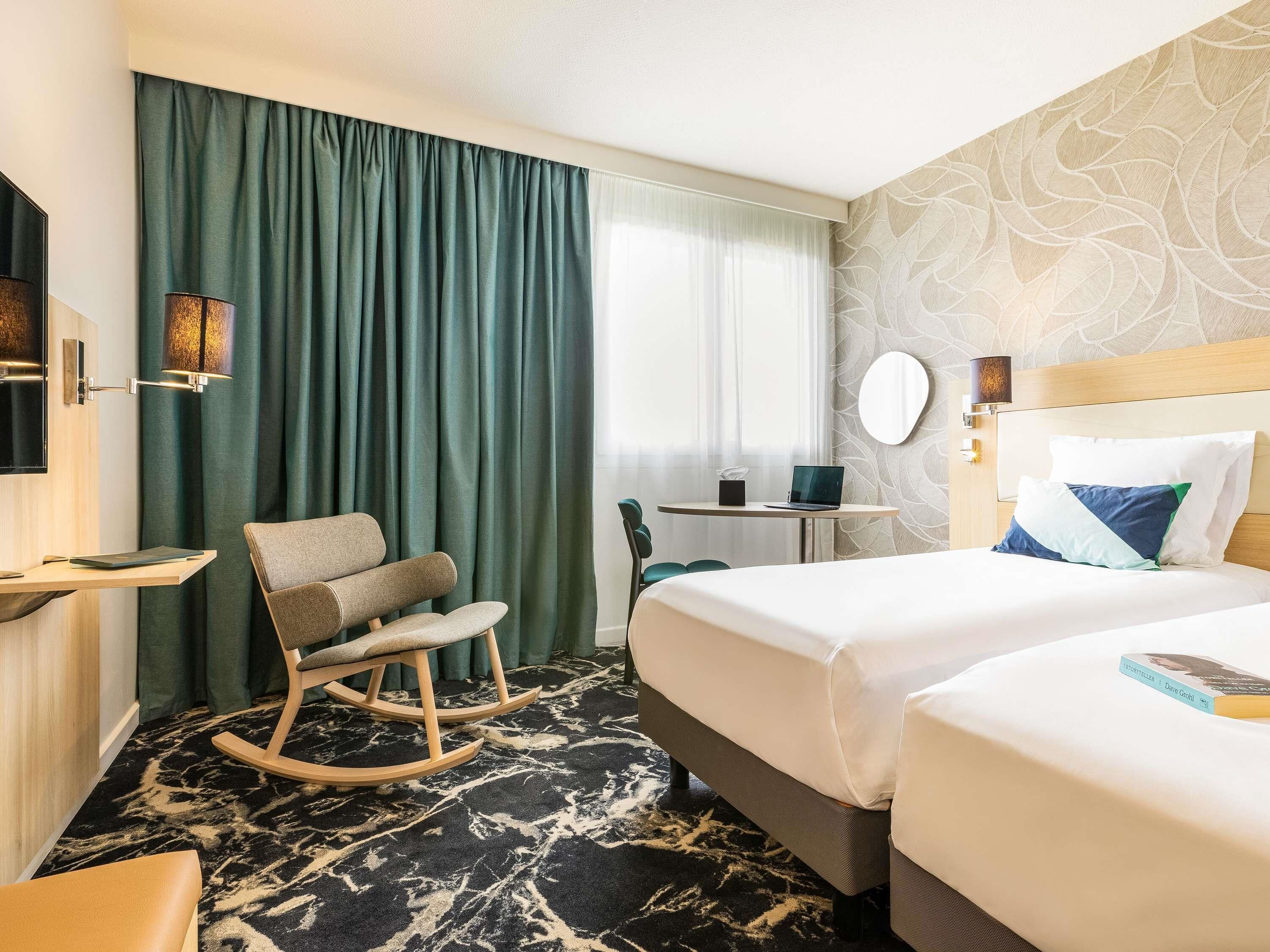 A hotel room with two beds, a desk, a chair, a lamp, and a window with curtains.