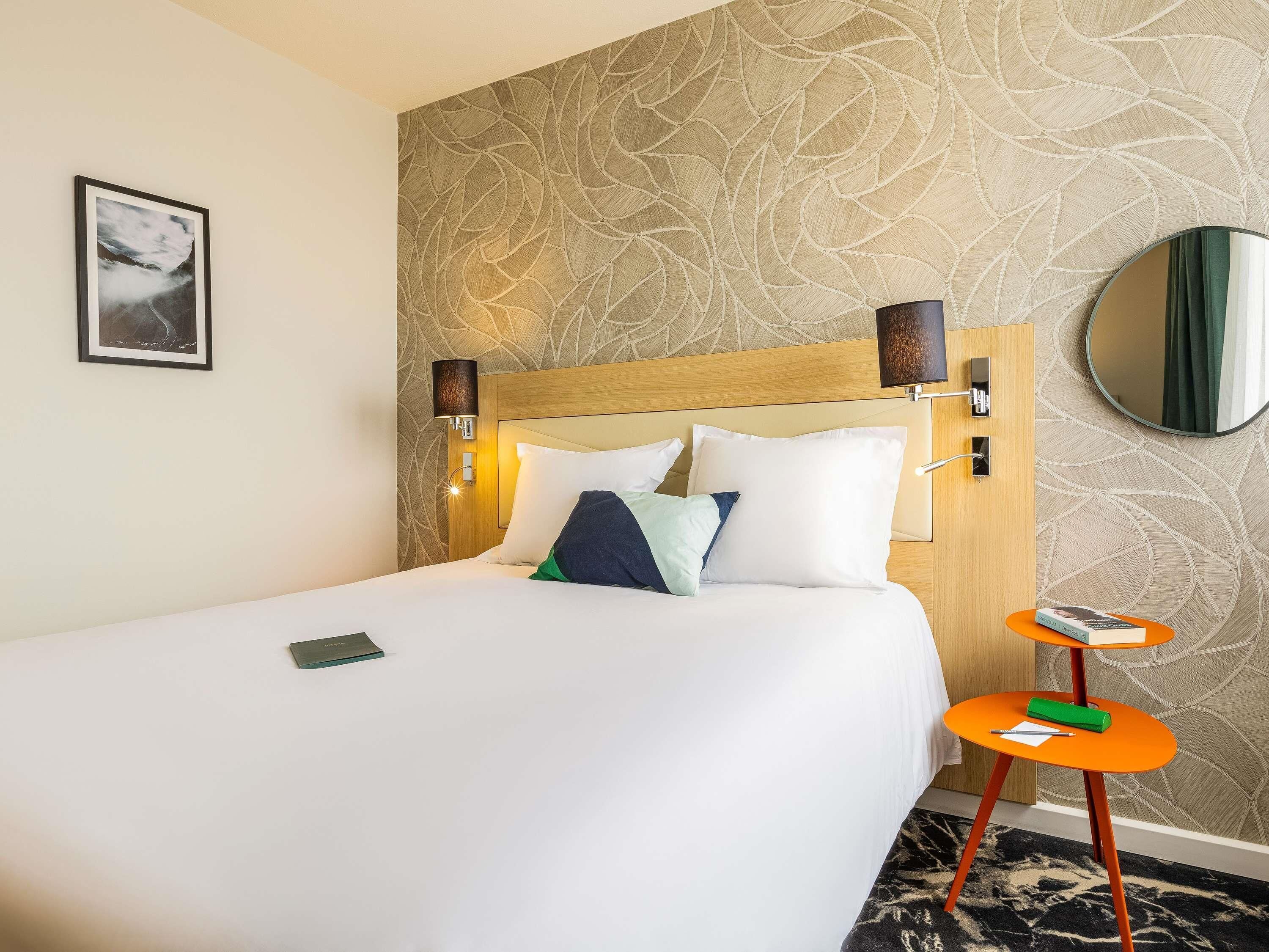 Une chambre d’hôtel avec un lit, une table de chevet, un miroir et un mur orné d’un motif de feuilles.