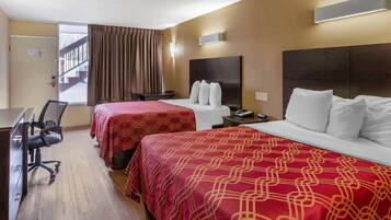 Ein Hotelzimmer mit zwei Betten, einem Schreibtisch und einem Stuhl. Ein Fenster mit Vorhängen, eine Wandleuchte und eine Tür sind zu sehen. Außerhalb der Tür ist eine Treppe sichtbar.