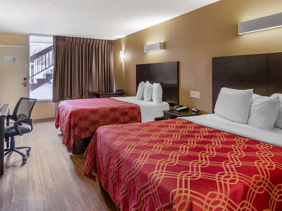 Ein Hotelzimmer mit zwei Betten, einem Schreibtisch und einem Stuhl. Ein Fenster mit Vorhängen, eine Wandleuchte und eine Tür sind zu sehen. Außerhalb der Tür ist eine Treppe sichtbar.