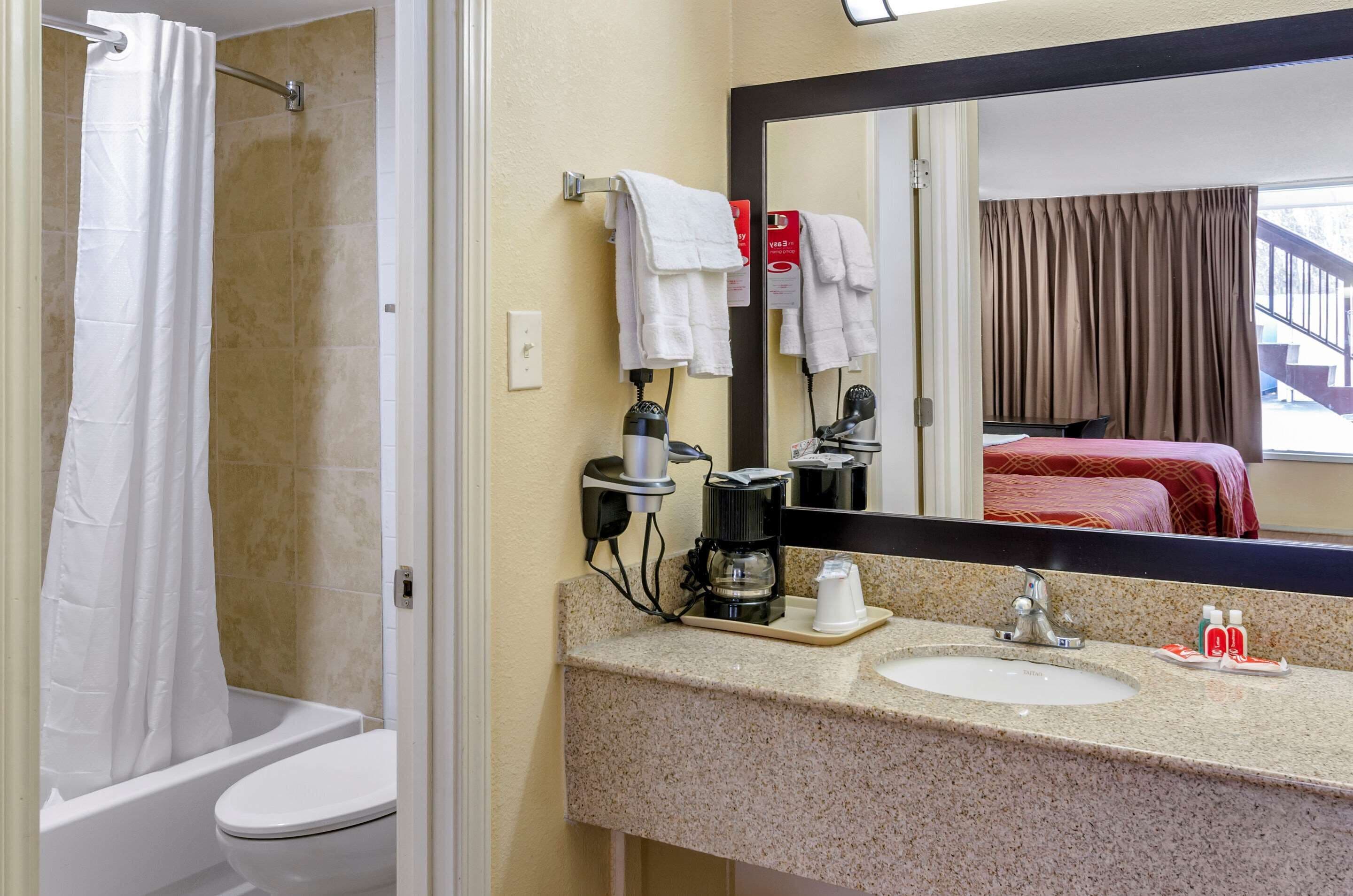 A bathroom with a shower, toilet, sink, and a mirror reflecting a bed and curtains.