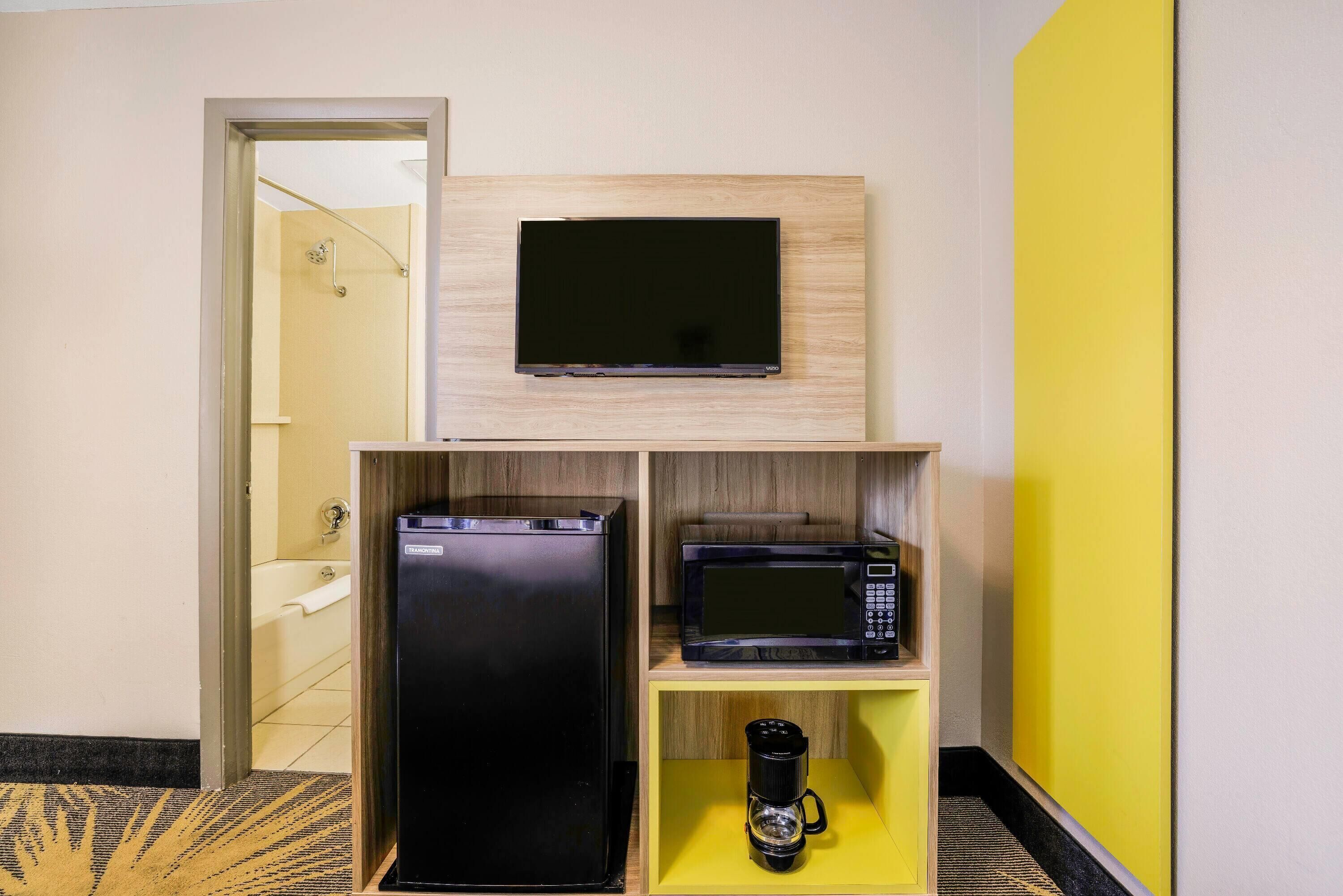 Habitación de hotel compacta con televisor de pantalla plana montado en la pared, un refrigerador negro, un microondas y una cafetera sobre un estante amarillo.
