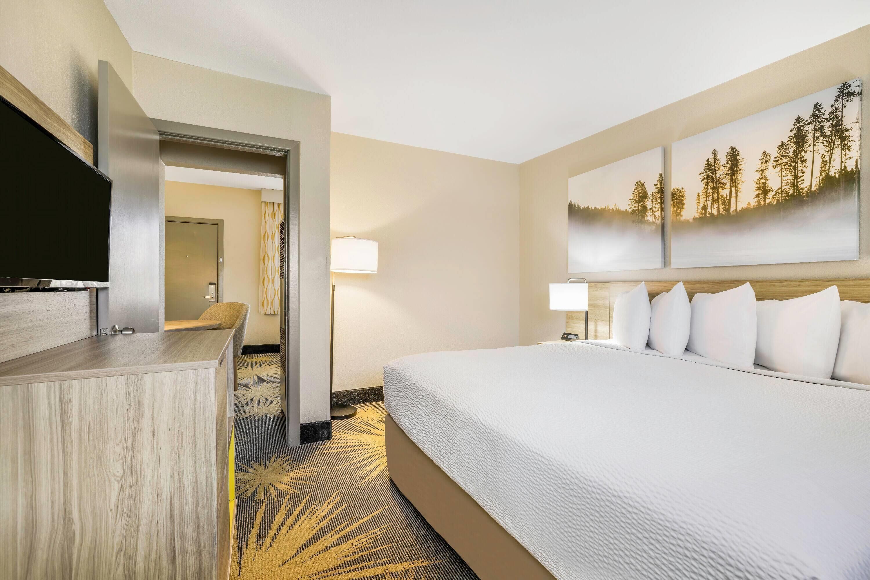 A hotel room with a large bed, a desk with a flat-screen TV, a wall-mounted TV, and a painting of trees.