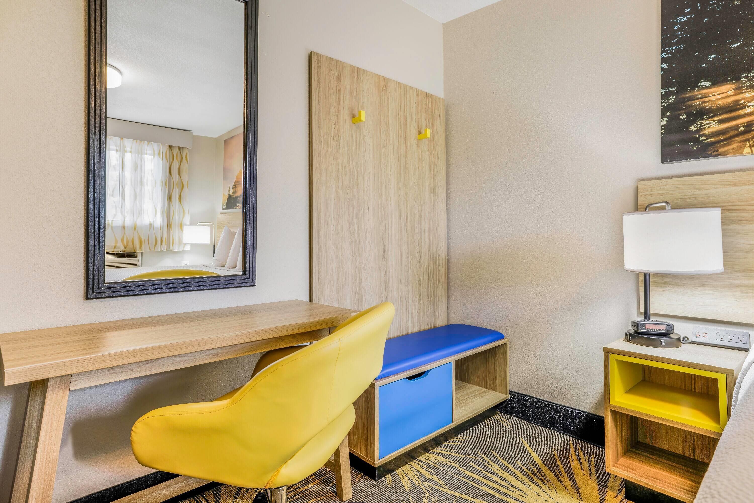 Een hotelkamer met een houten bureau, een gele stoel, een blauwe bank, een nachtkastje met een lamp en een kunstwerk aan de muur.
