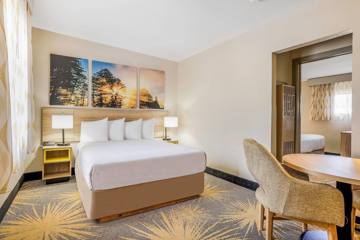 A hotel room with a large bed, two bedside tables with lamps, a dining area with a round table and chairs, and three wall-mounted paintings.