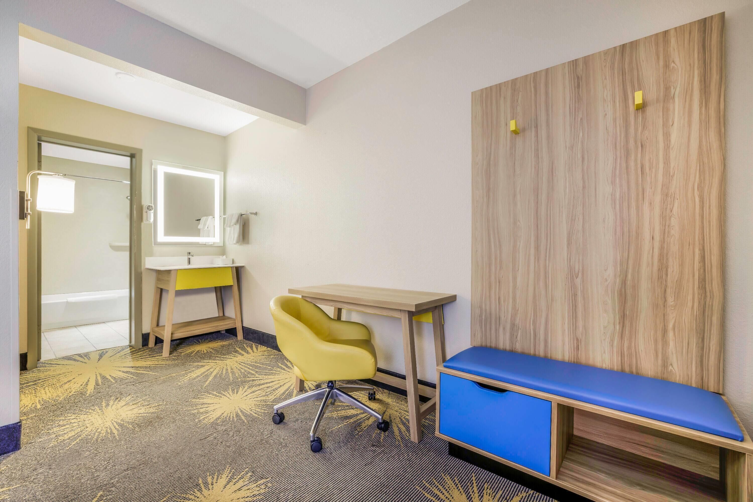 A hotel room with a desk, chair, and a bench.