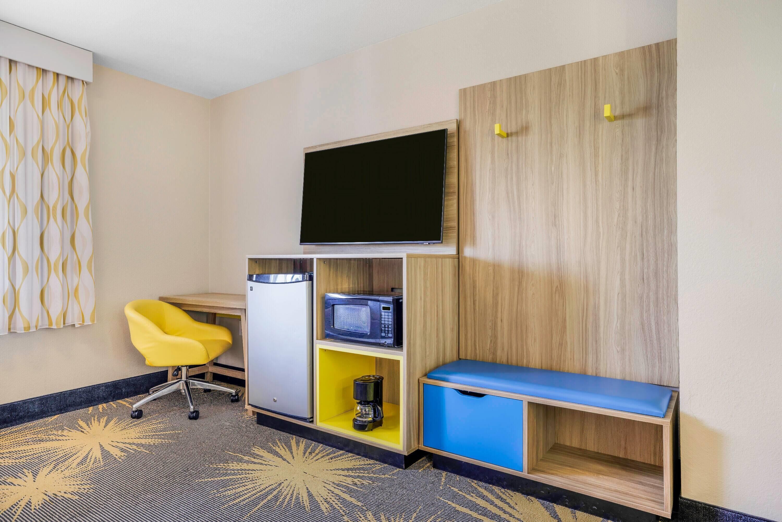 A hotel room with a flat-screen TV mounted on the wall, a yellow chair, a microwave, a coffee maker, and a blue bench.