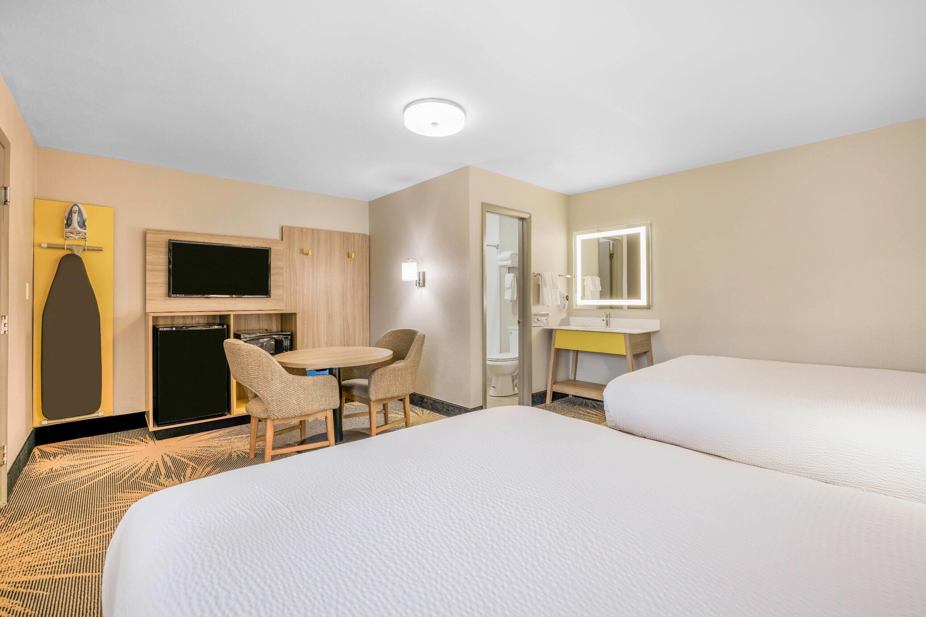 A hotel room with two beds, a desk with a chair, a TV, a small fridge, a mirror, and a bathroom visible through an open door.