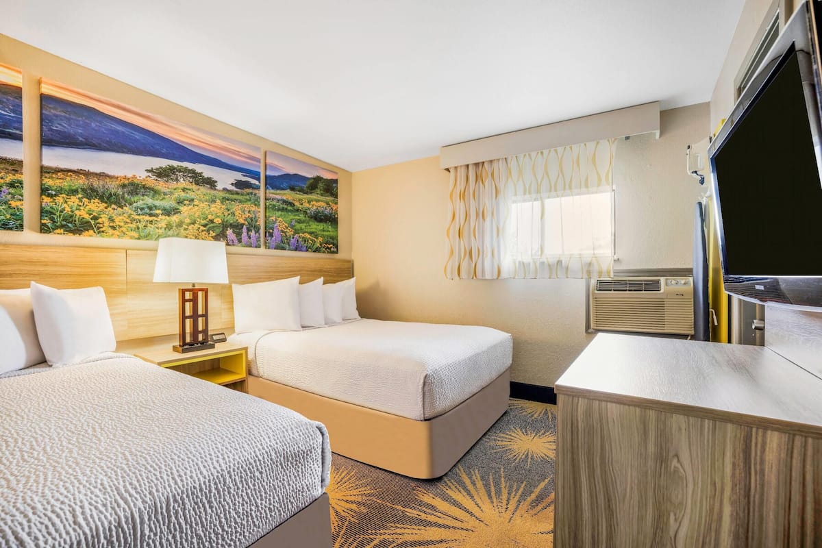 A hotel room with two beds, a desk, a TV, a painting, and an air conditioning unit.