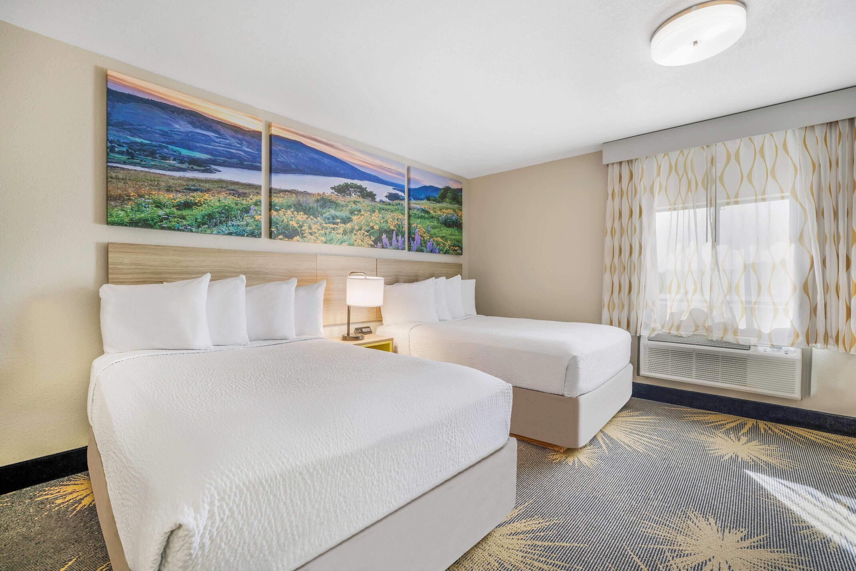 Una habitación de hotel con dos camas, un cuadro de paisaje grande sobre las camas, una lámpara en el techo y una ventana con cortinas estampadas.