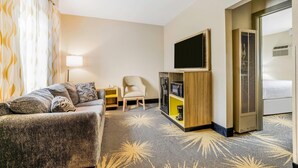 Ein Hotelzimmer mit einer Couch, einem Sessel, einem Fernseher, einer Minibar und einem großen Fenster mit Vorhängen.