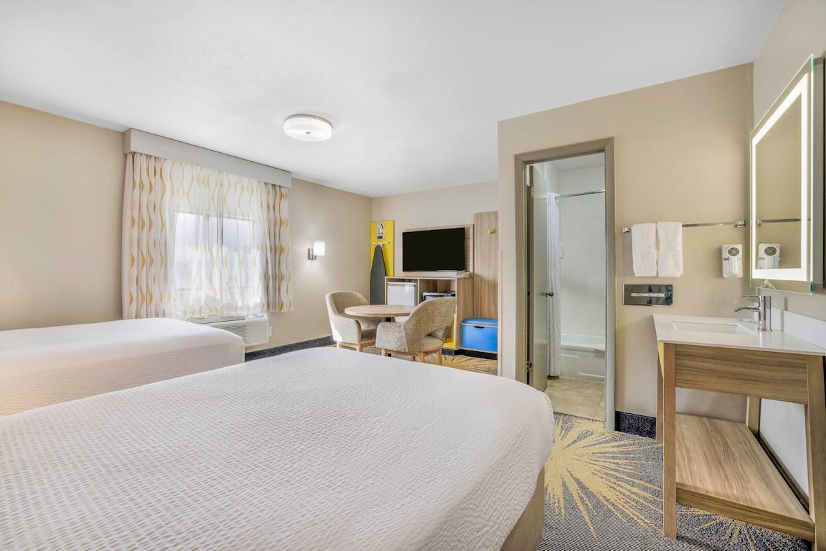 A hotel room with two beds, a desk with a chair, a TV, a bathroom with a mirror, and a small seating area.