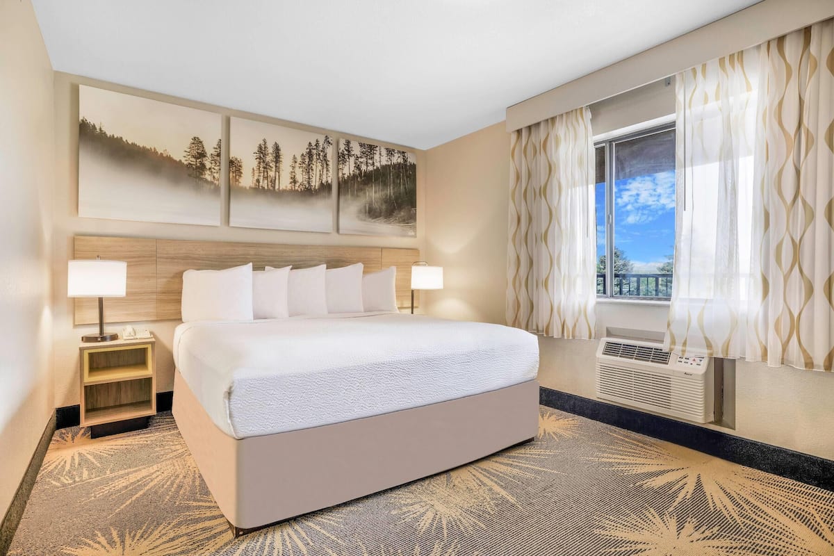 A hotel room with a large bed, two bedside tables with lamps, a wall-mounted air conditioner, and a window with curtains.