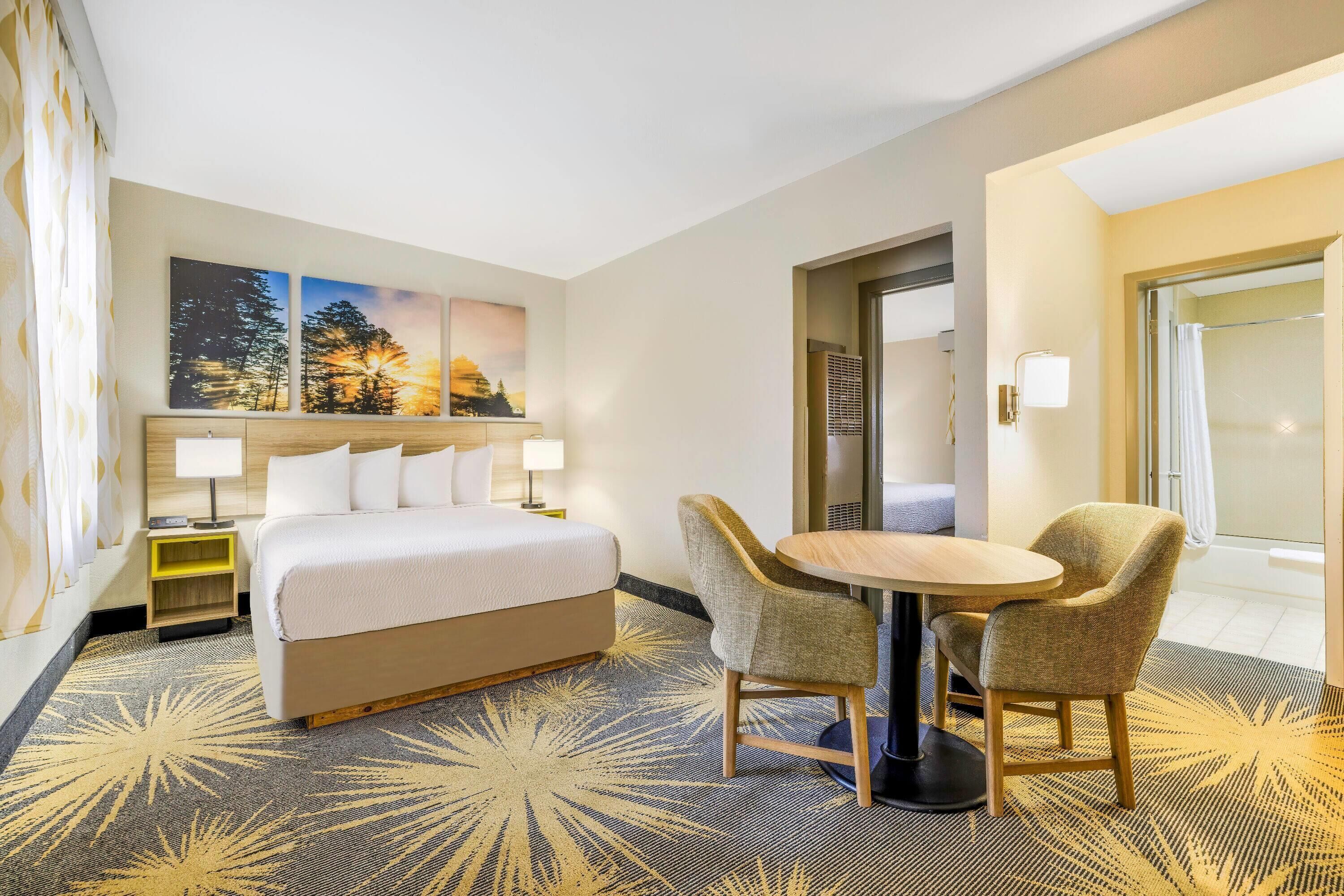 A hotel room with a bed, two chairs, a small table, a nightstand, and a wall-mounted lamp.