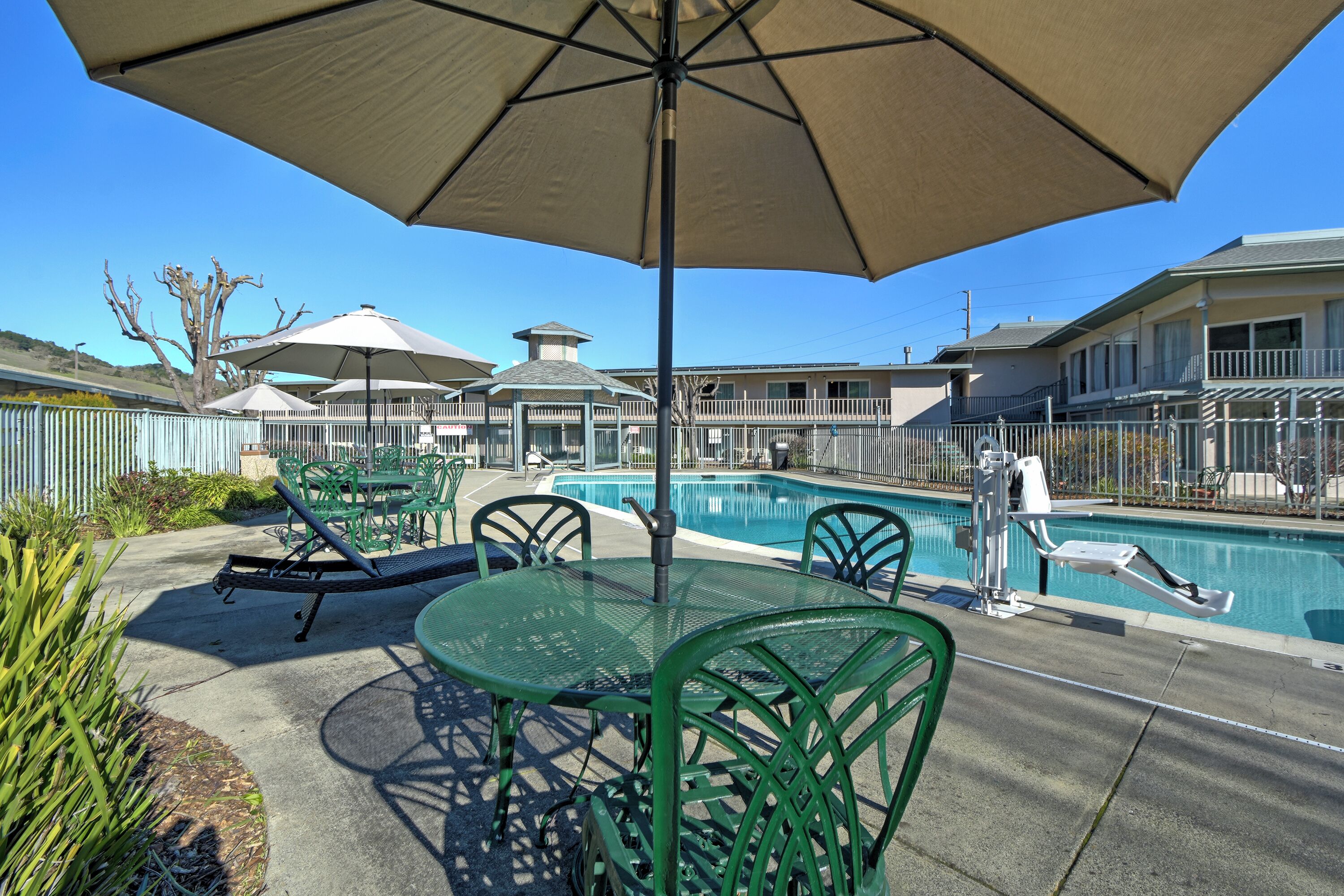 A pool area with a table and chairs, surrounded by umbrellas and a fence.