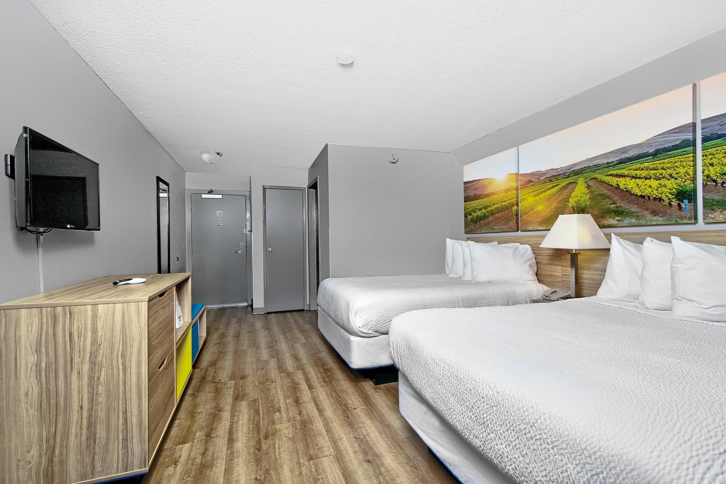 Une chambre d’hôtel avec deux lits, une télévision, un bureau, un miroir et un tableau représentant un vignoble.