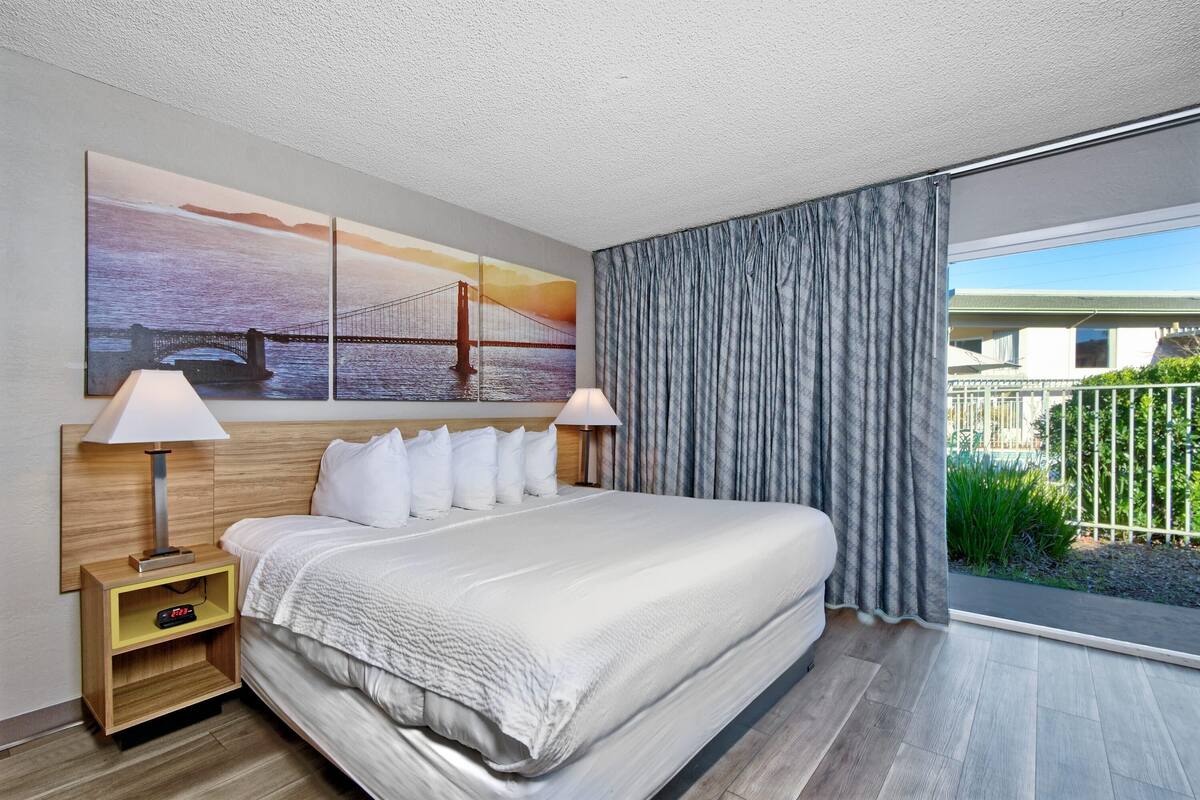 A hotel room with a large bed, two bedside tables with lamps, a wall-mounted artwork of a bridge, and a view of a building through a window with curtains.