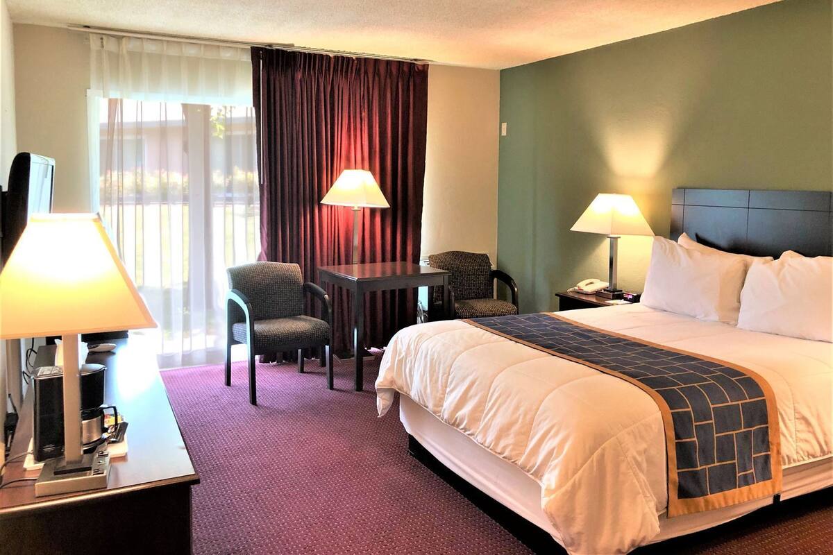 A hotel room with a bed, two chairs, a desk with a lamp, a telephone, and a window with curtains.