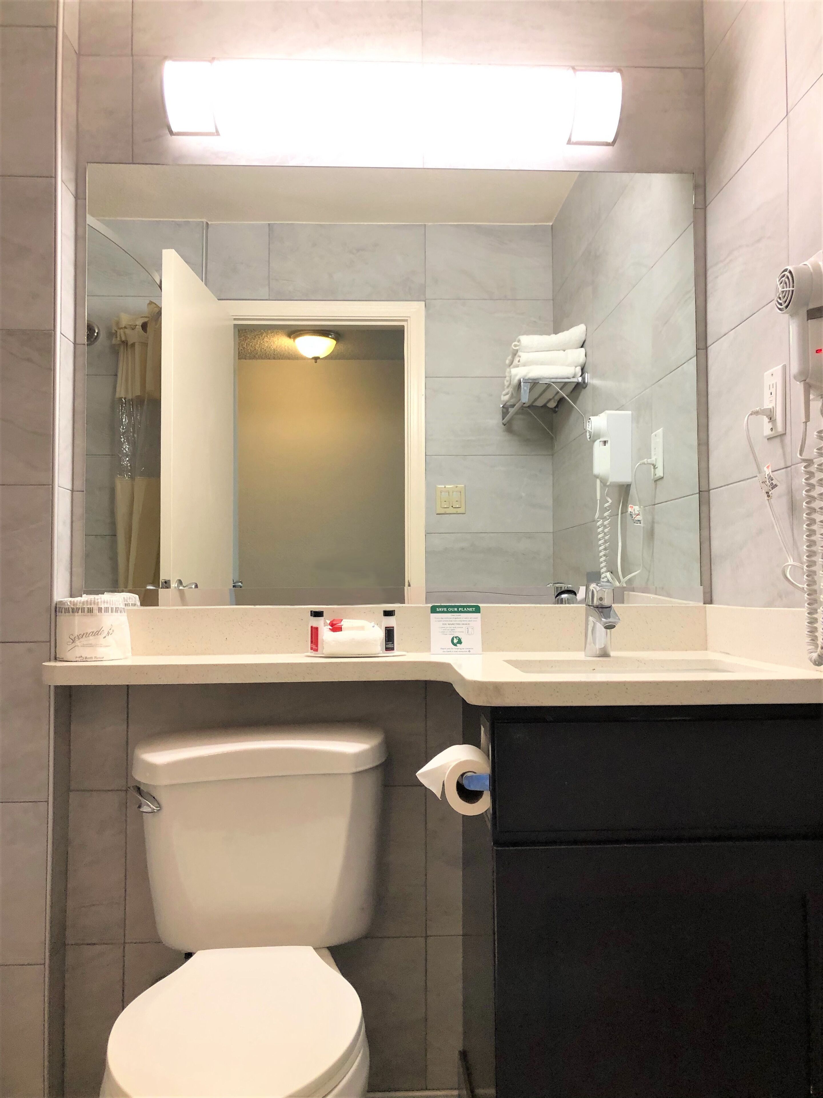 Ein Badezimmer mit Toilette, Waschbecken und Spiegel. Ein Handtuchhalter mit Handtüchern, ein Handtrockner und ein Schild zur Förderung des Umweltschutzes sind vorhanden.