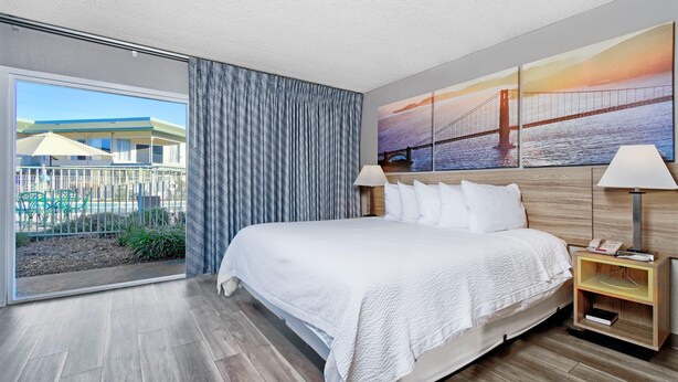 A hotel room with a large bed, a nightstand with a lamp, a painting of a bridge, and a view of a building and greenery through the window. - Days Inn by Wyndham Novato/San Francisco (Novato)