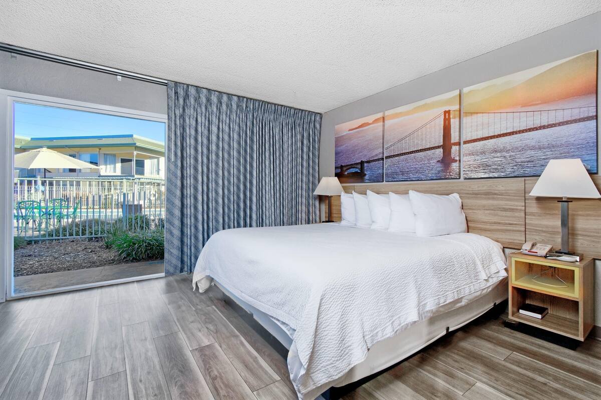 A hotel room with a large bed, a nightstand with a lamp, a painting of a bridge, and a view of a building and greenery through the window.