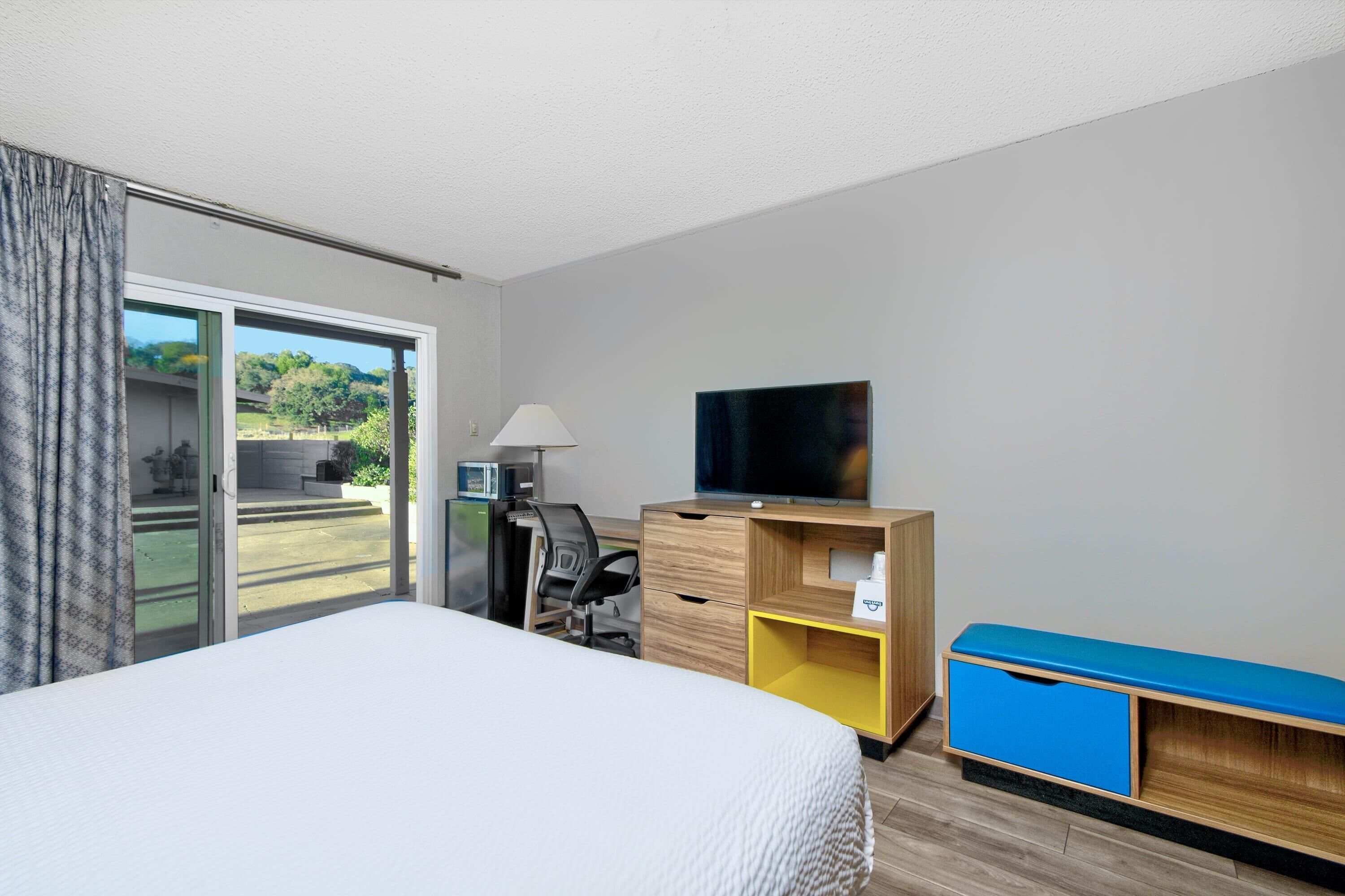 A hotel room with a bed, a desk with a chair, a television, and a sliding door to the outside.