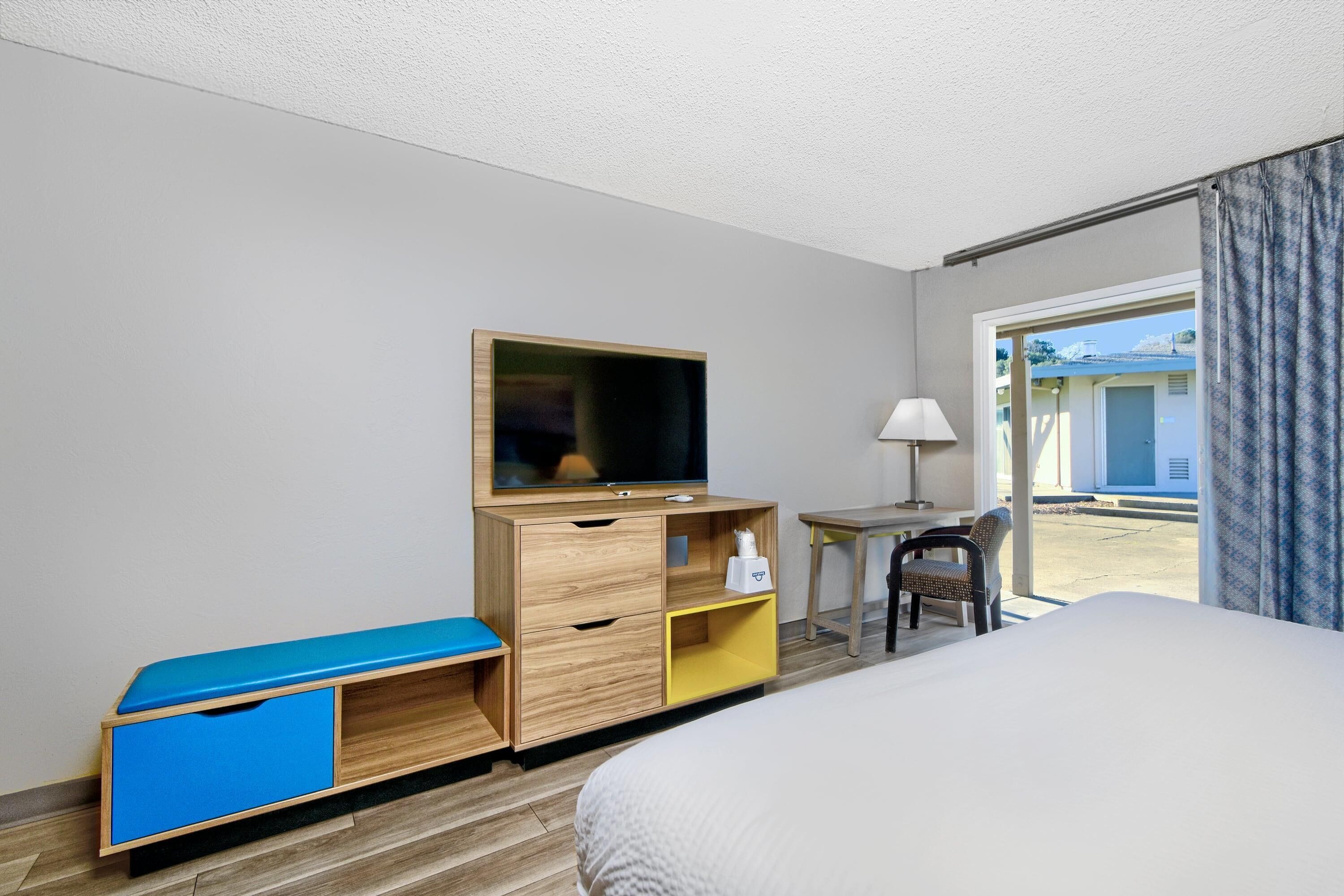 A hotel room with a bed, a TV on a wooden dresser, a desk with a chair, and a view of the outside.