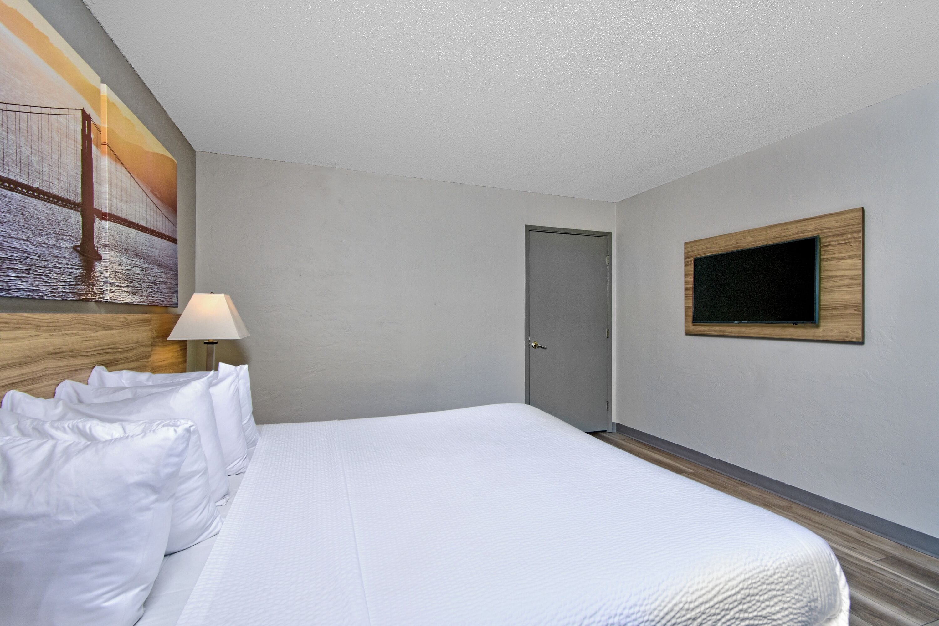 A hotel room with a large bed, a door, a wall-mounted TV, and a painting on the wall.