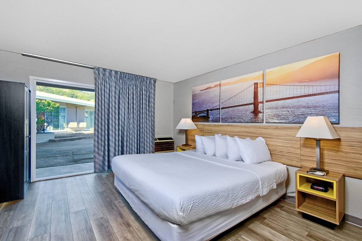 A modern hotel room with a large bed, two bedside tables, a nightstand, and a view of a bridge through the window.