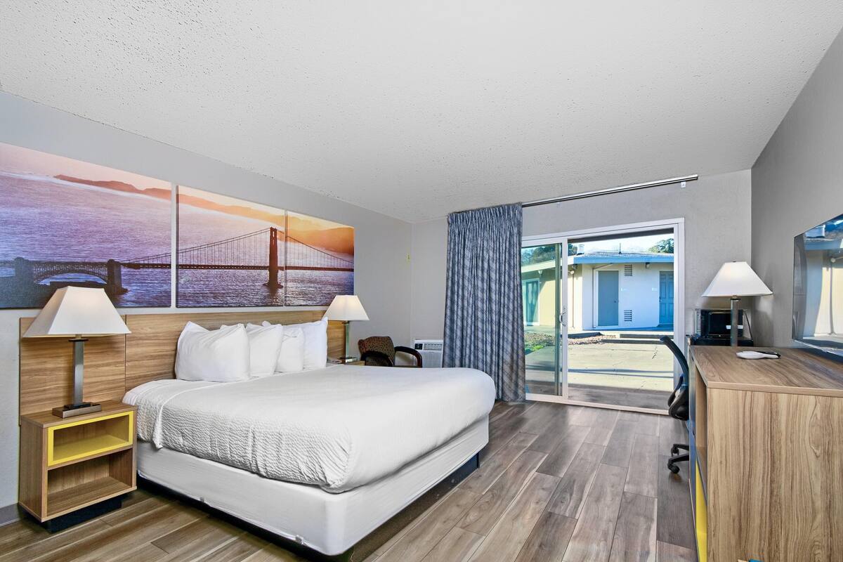 A hotel room with a large bed, two bedside tables with lamps, a desk, and a view of the outside through a glass door.