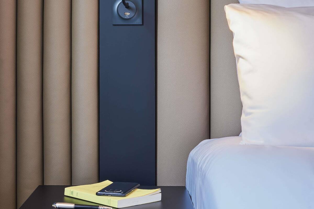 A neatly made bed with a pillow, a bedside table with a book and a pen, and a power outlet with a cable plugged in.