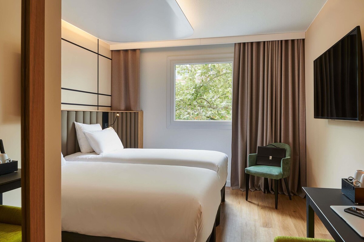 Habitación de hotel con dos camas, un escritorio, una silla, un televisor y un ventanal con cortinas.