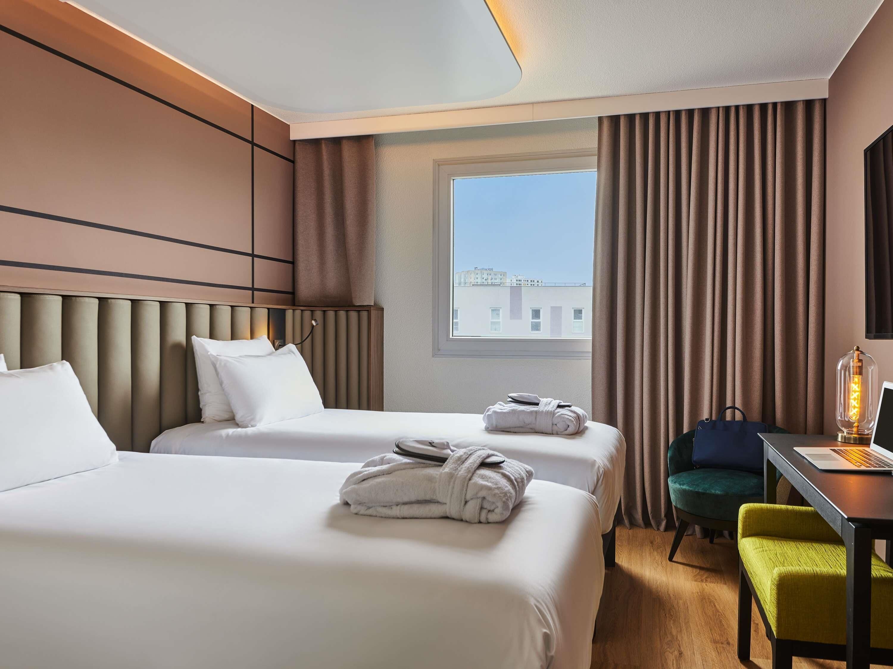 A hotel room with two beds, a desk with a laptop, a chair, and a lamp.