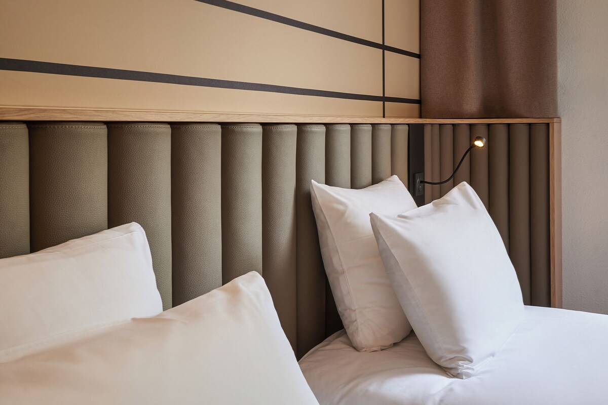A modern hotel room with a padded headboard, white bedding, and a neutral color scheme.
