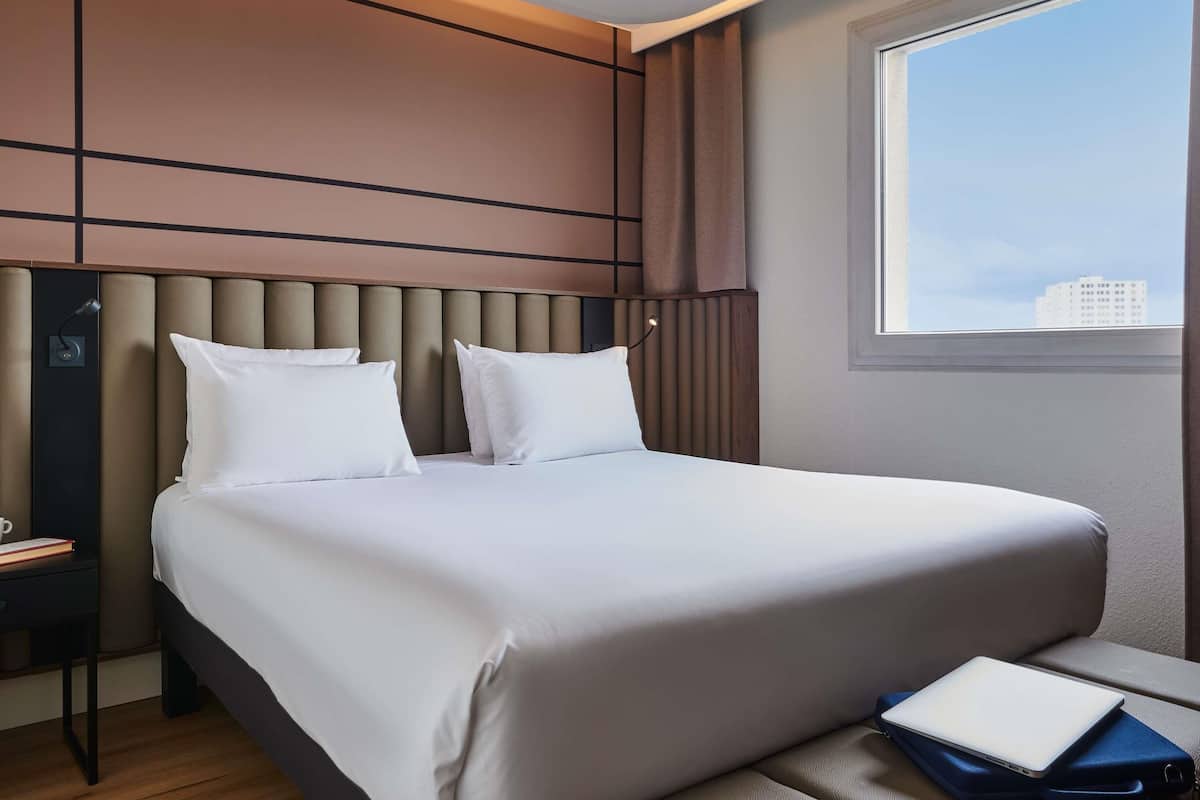 A modern hotel room with a large bed, a bedside table with a lamp, and a view of the city through a window.
