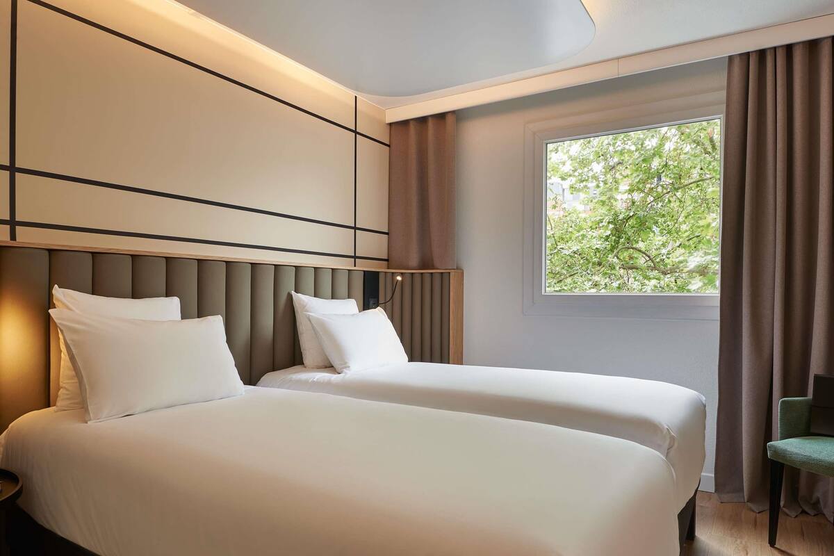 A hotel room with two beds, a large window with curtains, and a view of greenery outside.