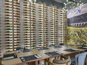 Ein Restaurant mit einer Weinschlange, einem langen Esstisch gedeckt mit Gläsern und Besteck, und einem Wandgemälde eines Weinbergs.