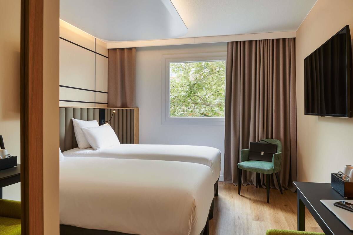 A hotel room with two beds, a desk, a chair, a TV, and a window with curtains.