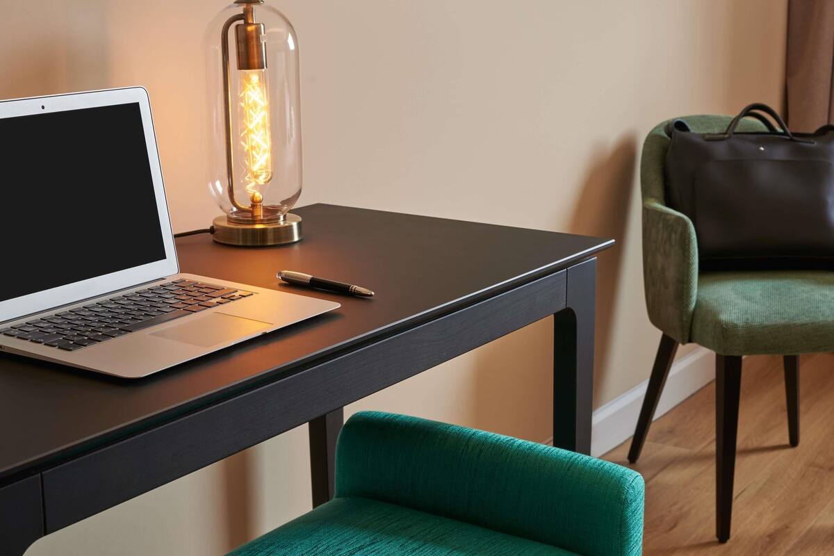 A laptop on a desk with a vintage-style lamp, a green chair, and a leather bag.