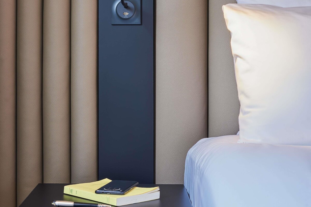 A neatly made bed with a pillow, a bedside table with a book and a pen, and a power outlet with a cable plugged in.