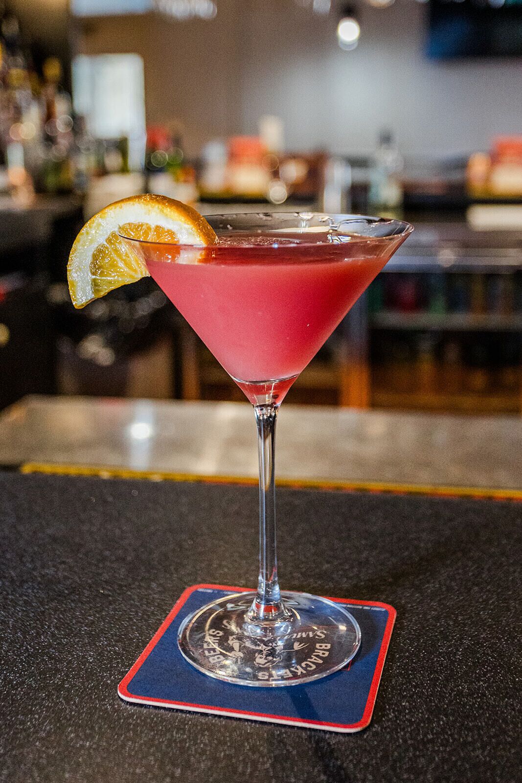 A cocktail in a martini glass with a red rim, garnished with an orange slice, on a coaster with a blue and red design.