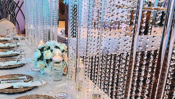 Uma longa mesa de banquete posta com pratos dourados ornamentados, guardanapos brancos e copos de cristal, rodeada por decorações de cristal penduradas.