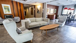Um saguão de hotel moderno com um amplo sofá bege, duas poltronas, uma mesa de centro de madeira e uma área de refeições com luminárias pendentes vermelhas.
