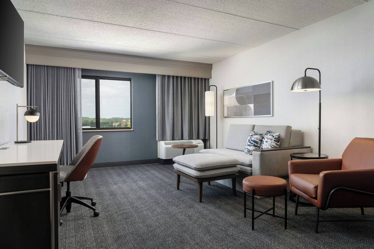 A hotel room with a desk, two chairs, a sofa, a small table, and a floor lamp. There is a window with curtains, a wall-mounted air conditioner, and a framed abstract artwork.