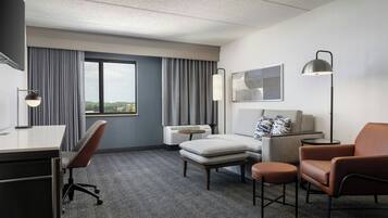 A hotel room with a desk, two chairs, a sofa, a small table, and a floor lamp. There is a window with curtains, a wall-mounted air conditioner, and a framed abstract artwork.