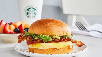 A breakfast sandwich with bacon, lettuce, and egg on a bun, a plate of fruit, and a Starbucks cup in the background.