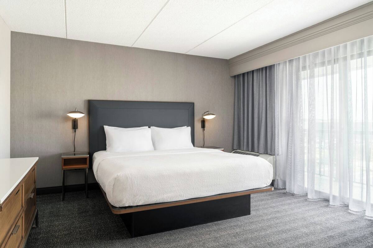 A hotel room with a large bed, two bedside lamps, a nightstand, and a view of the outdoors through sheer curtains.