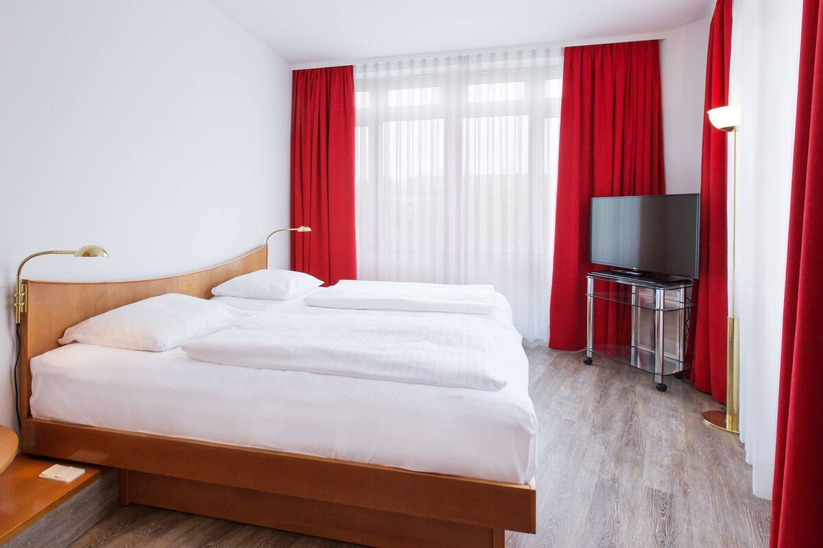A hotel room with a wooden bed, white bedding, a TV on a stand, and red curtains.