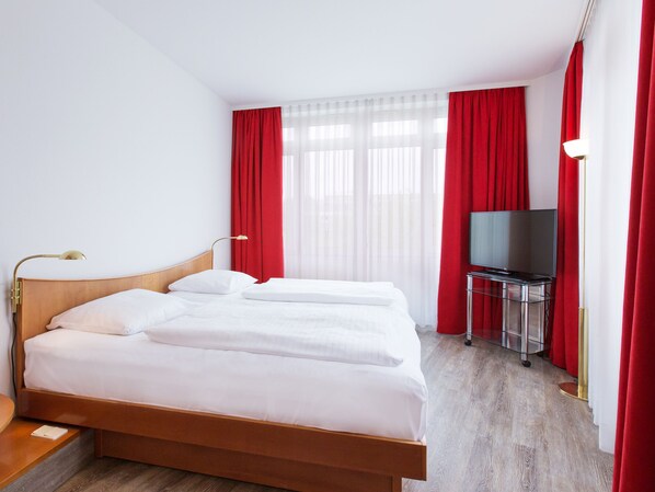 Una habitación de hotel con una cama de madera, ropa de cama blanca, un televisor sobre un pedestal y cortinas rojas.