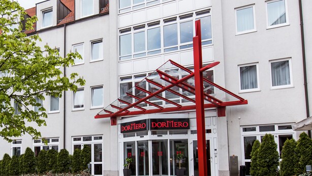 Un edificio moderno con un toldo rojo sobre la entrada y el nombre 'DORMERO' exhibido sobre las puertas.